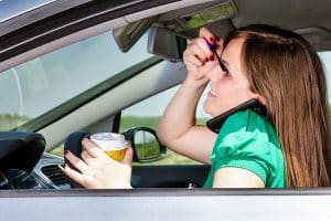 Pretty young woman applying makeup, speaking on phone and drinking coffee while driving her car